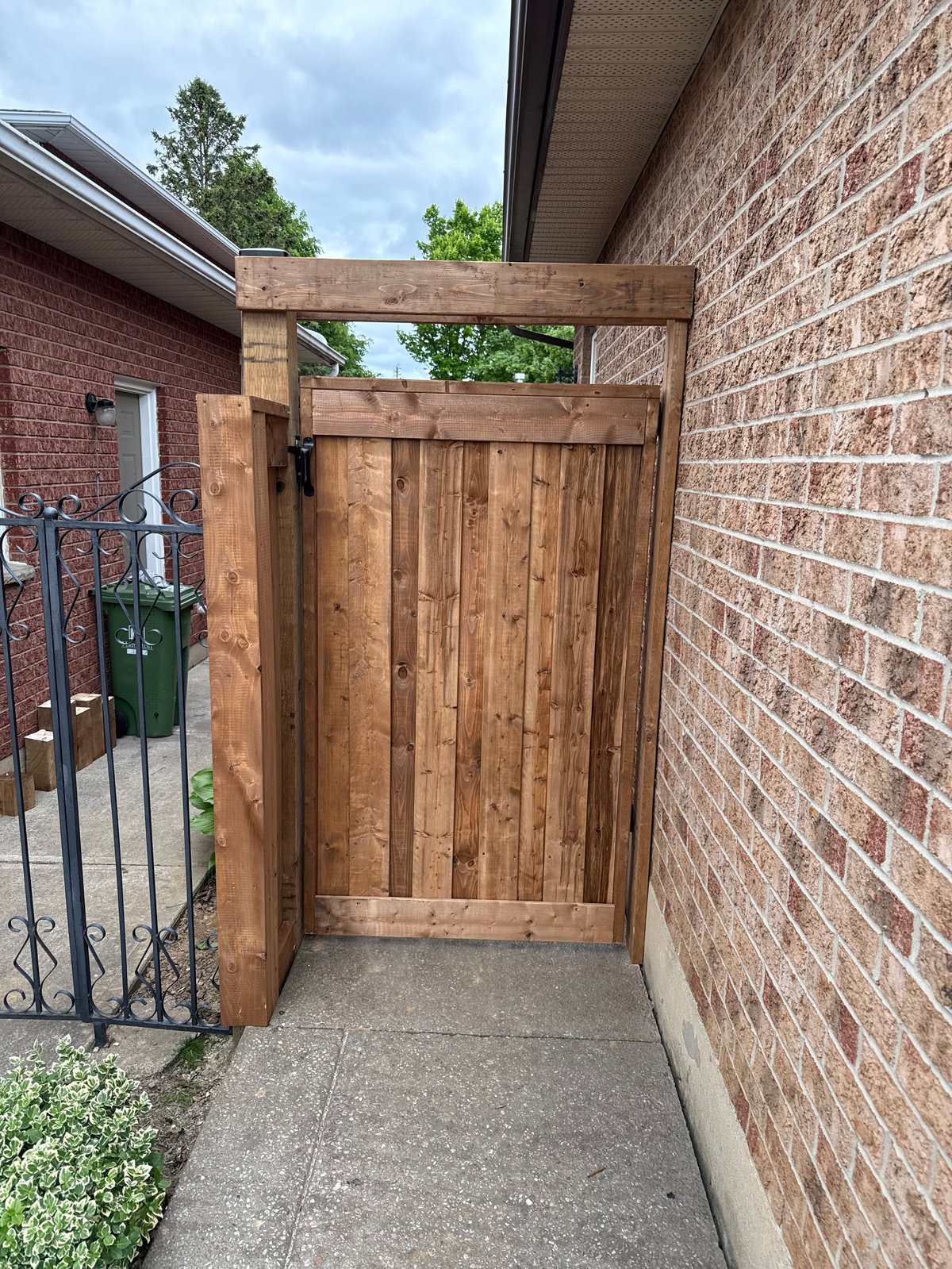 Wooden gate with pergola top attached to brick house and metal fence, leading to a concrete pathway