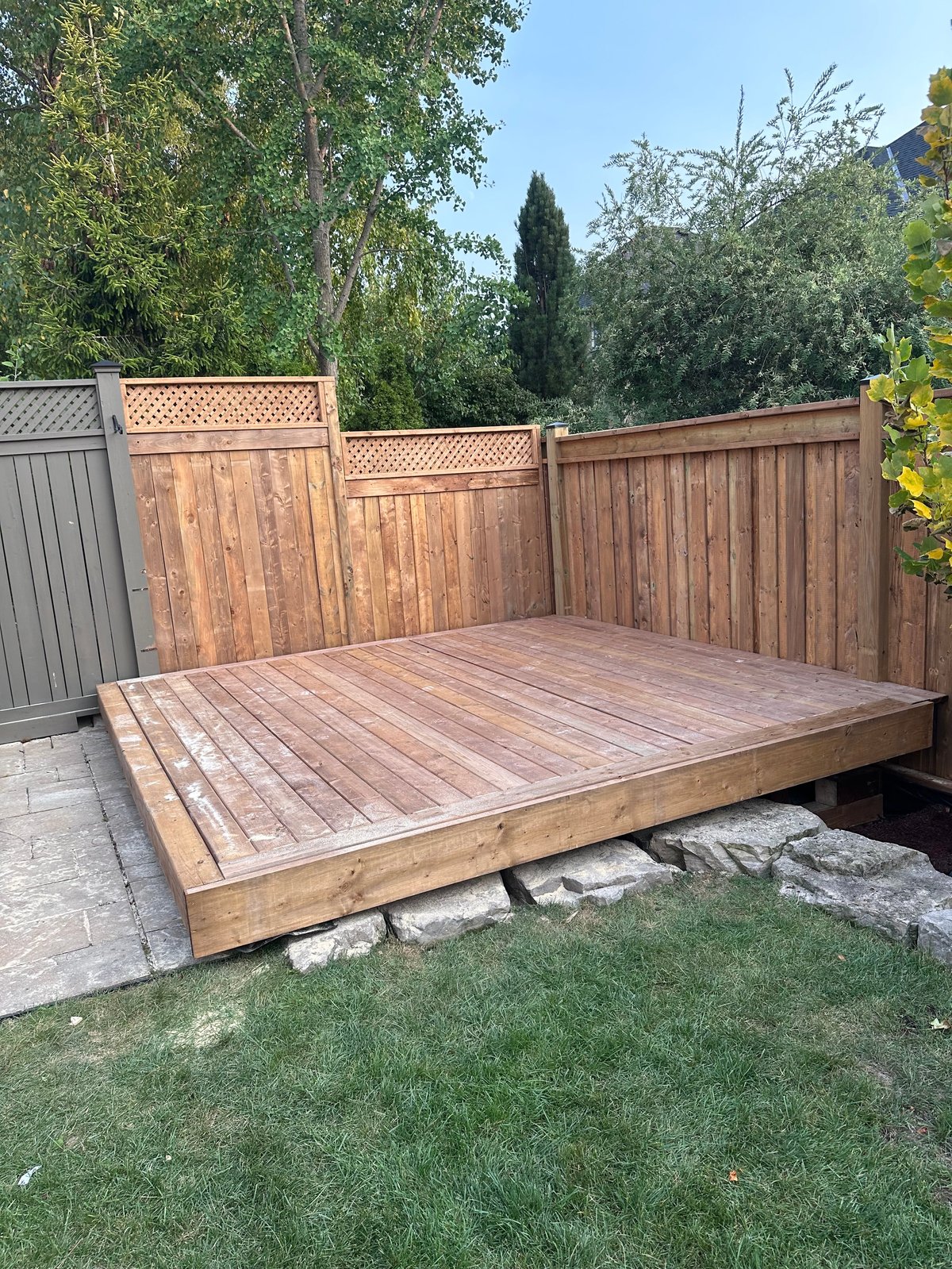 Wooden elevated deck with lattice fence in backyard surrounded by green lawn and mature trees