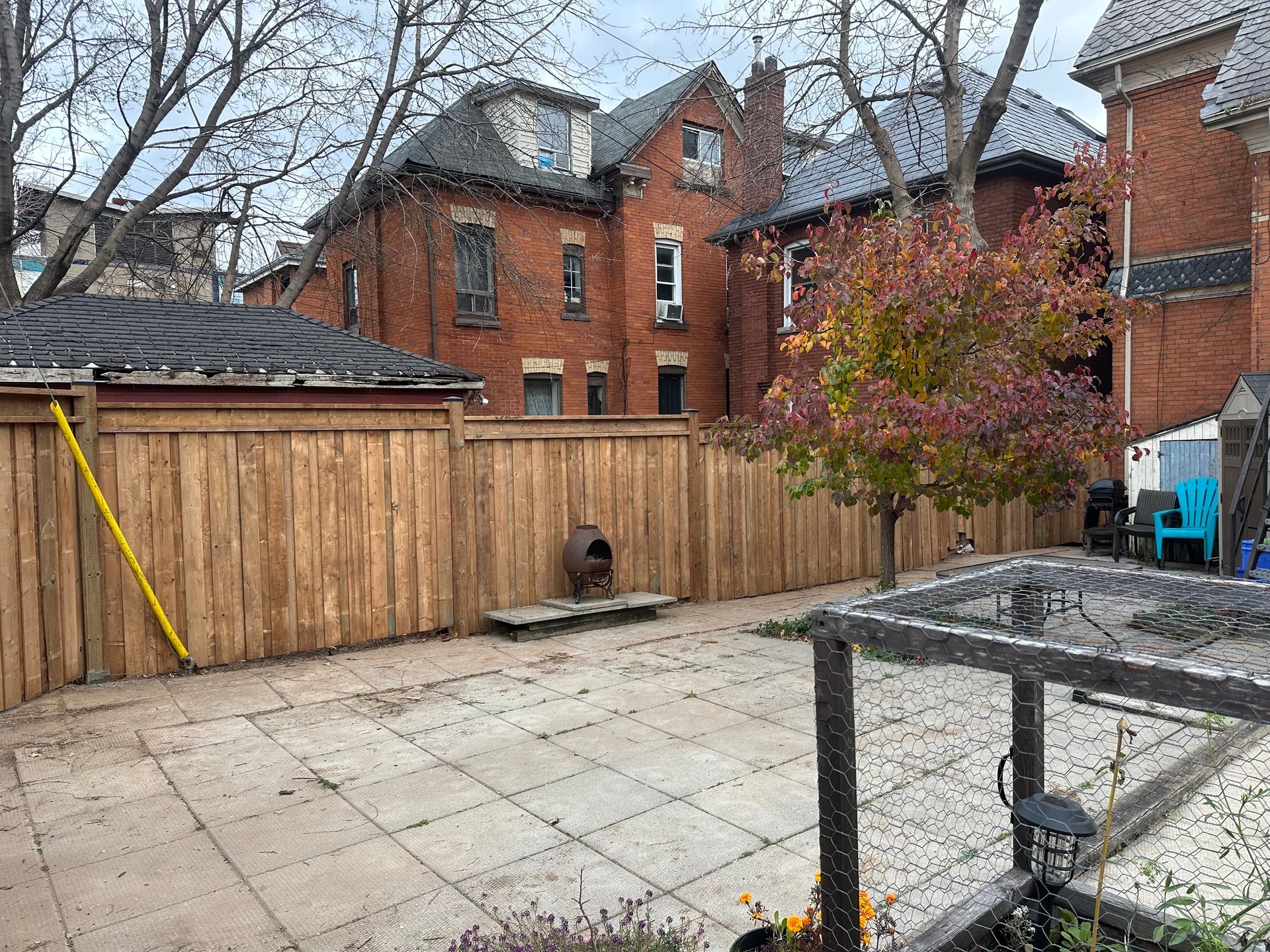 Patio area with paving stones enclosed by wooden fence, featuring a picnic table in foreground and brick Victorian house with bare trees in background