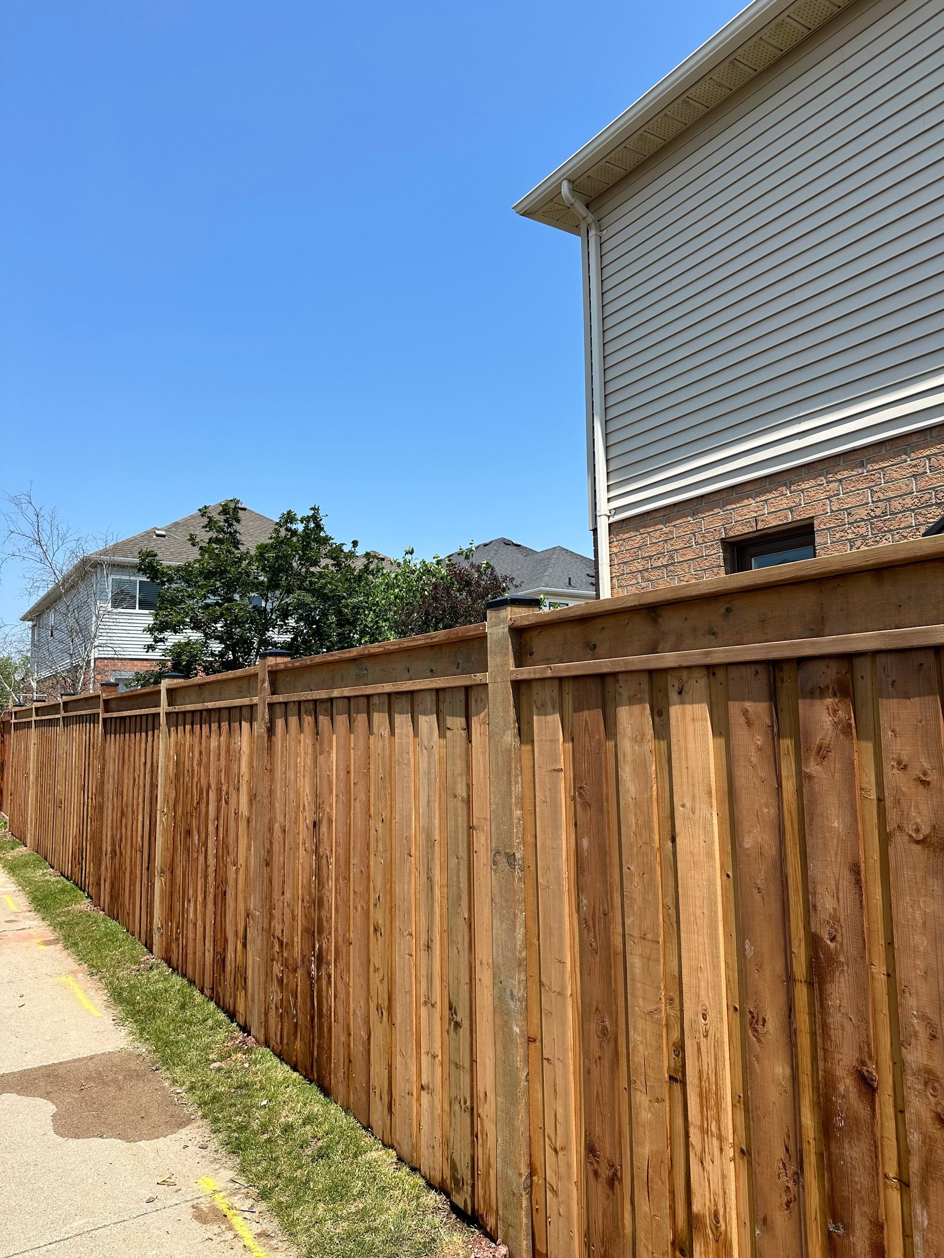 Wooden fence along residential property with house and mountains visible in background under clear blue sky